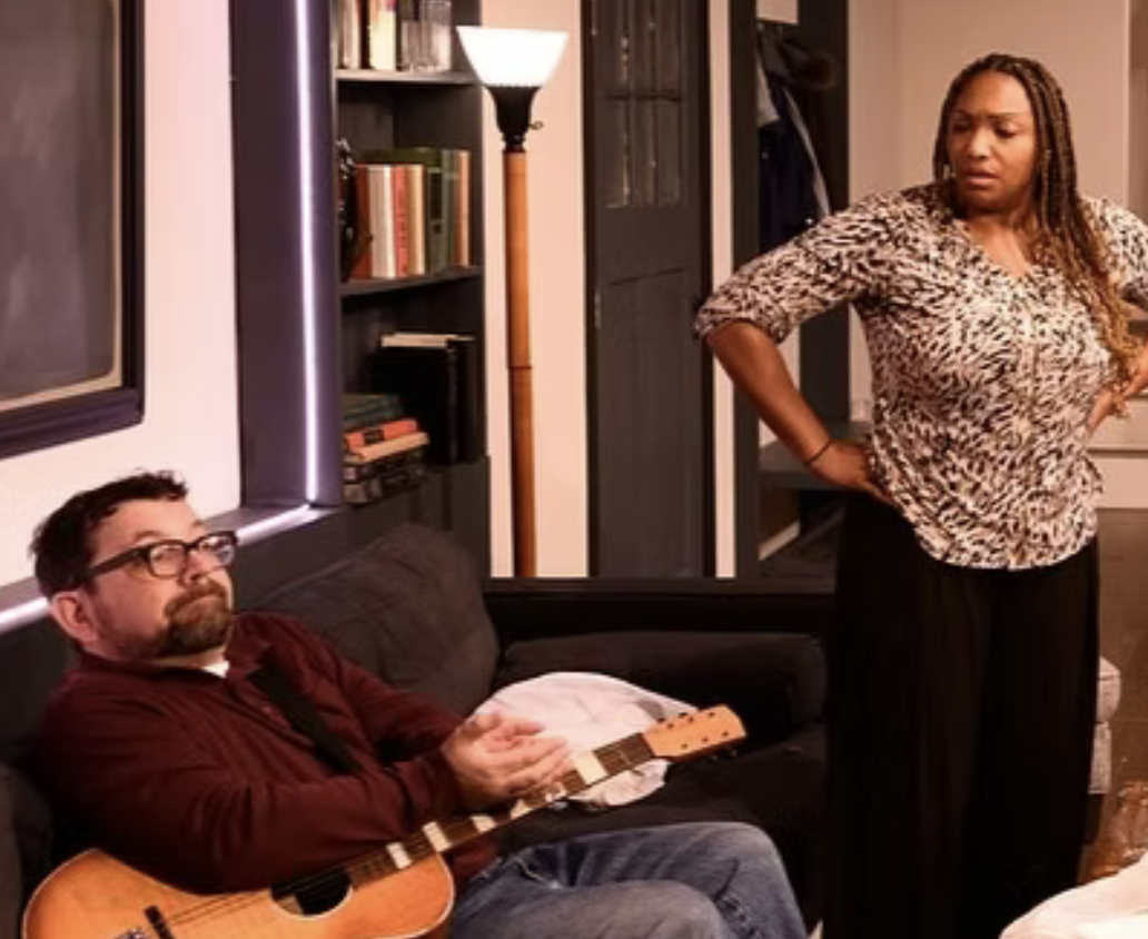 Production photo of Redtwist production of How to Transcend a Happy Marriage by Sarah Ruhl, directed by Elizabeth Swanson. An upper middle class living room. A middle aged white man sits on a couch with a guitar and holding something tiny and fragile in his hands. A middle aged Black woman looks on in emotional distress.